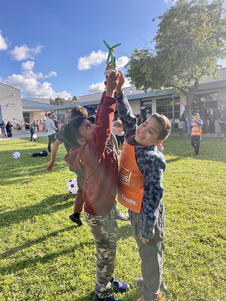"Two boys hold up a small wind turbine they built together at an outdoor school event, with one boy wearing a Home Depot Kids Workshop apron."