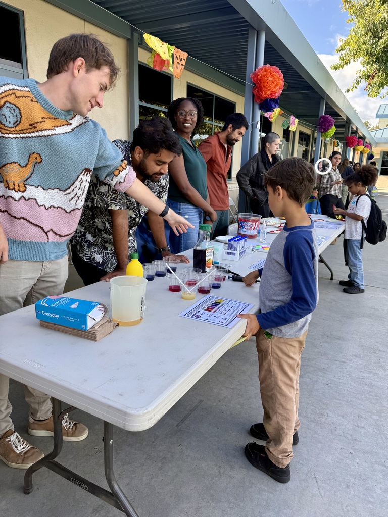 "University students demonstrate a colorful liquid science experiment to a young child at an outdoor table, with cups of colored liquids and a pH chart visible."