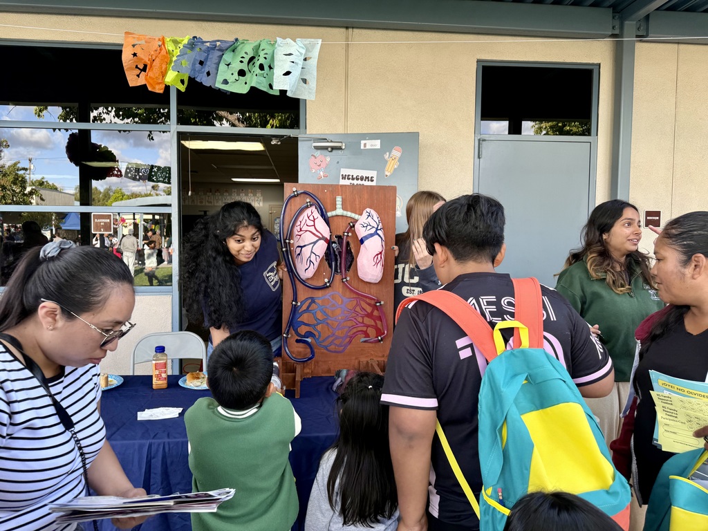  "A student presenter shows a model of the human heart and lungs to a group of children and adults at an outdoor school event booth, decorated with colorful papel picado."