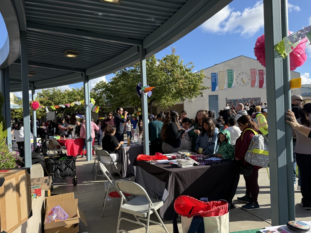 "Families and community members browse vendor and information tables at an outdoor school event, decorated with colorful paper flowers and papel picado banners."