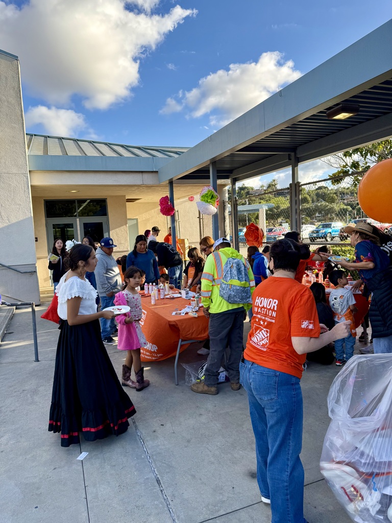 "Home Depot volunteers in orange shirts assist families at a craft activity table at an outdoor school event, with a girl in traditional folklórico dress visible in the foreground."