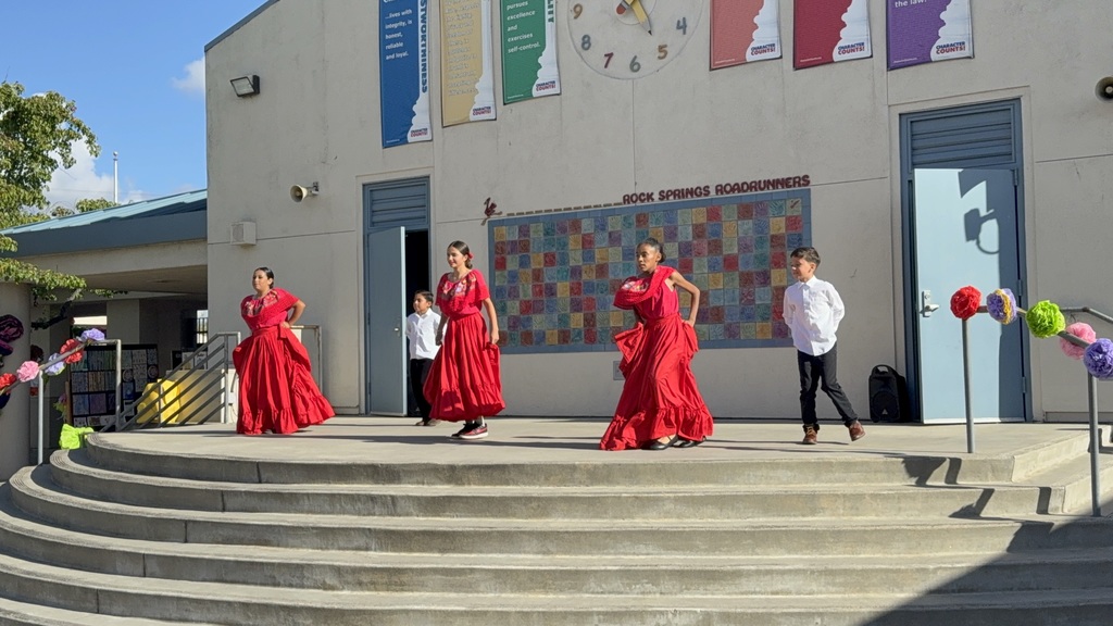 "Rock Springs Ballet folklorico dancers mid-performance, holding the edges of their large, colorful skirts aloft to create a fan-like effect while dancing at a school community event."