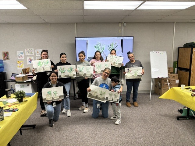 Six moms, a child, and two family liaisons pose for a picture inside room 21 while holding up their artwork