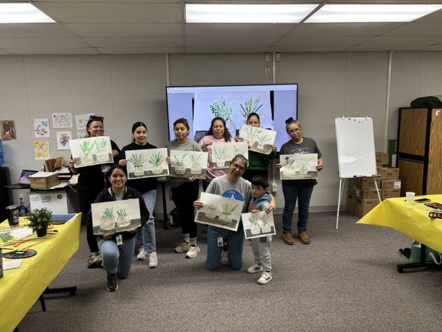 Six moms, a child, and two family liaisons pose for a picture inside room 21 while holding up their artwork