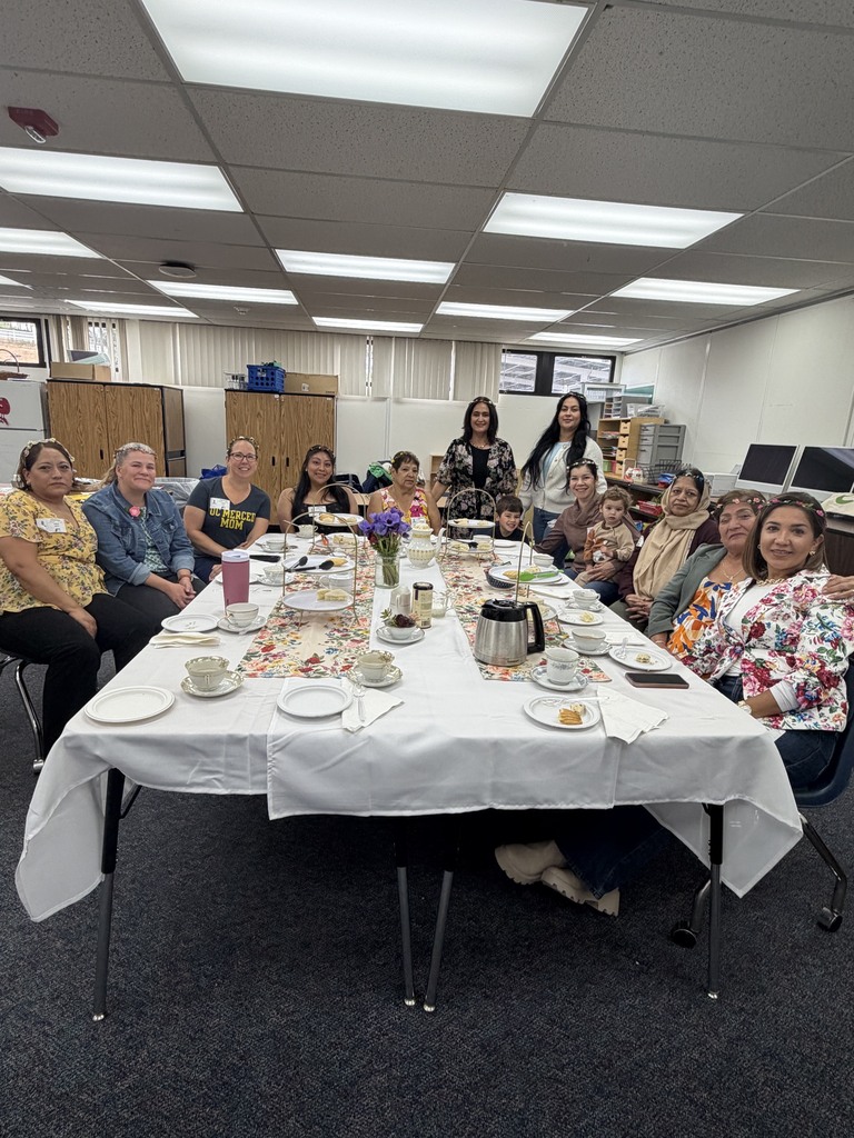 A group of women seated around a long, decorated tea party table with floral runners, teacups, and treats.