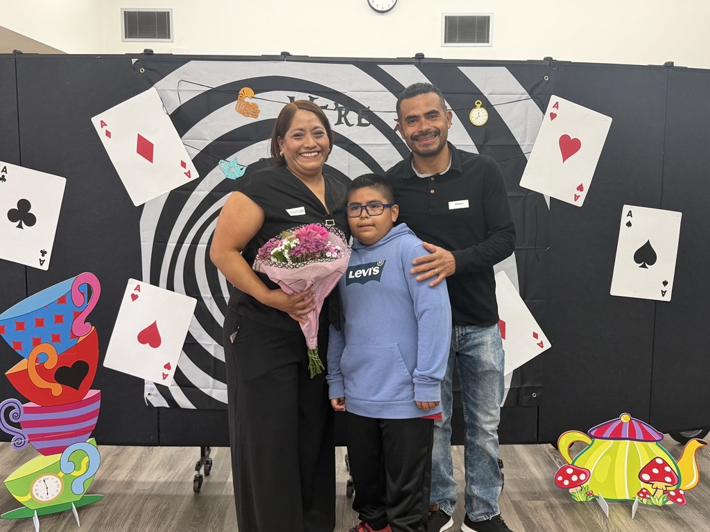 In image IMG_2684.jpg, a smiling family of three poses together by an Alice in Wonderland themed photo backdrop