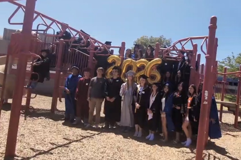 Graduating seniors gather on a playground structure around gold “2026” balloons for a group photo.