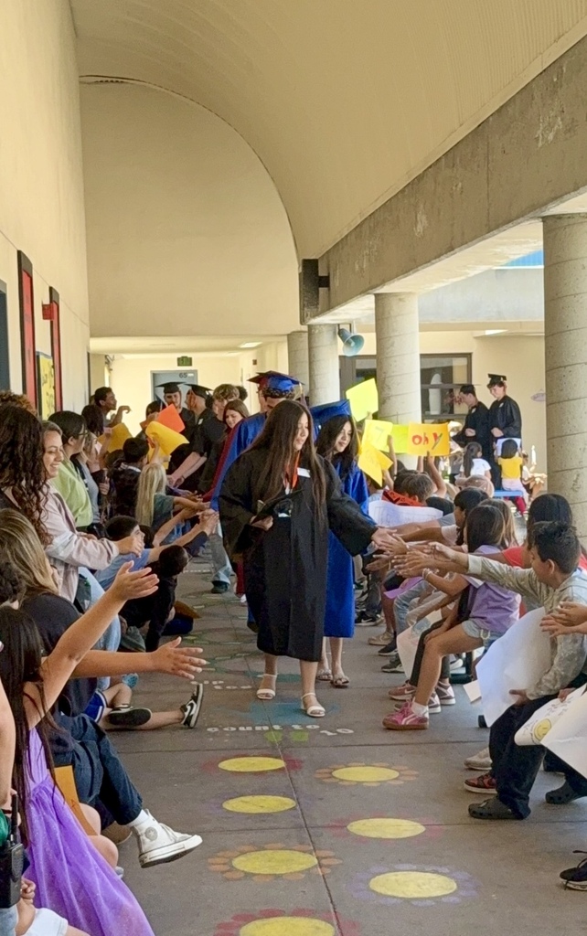 Students line a school walkway as graduating seniors in caps and gowns walk through, giving high-fives.