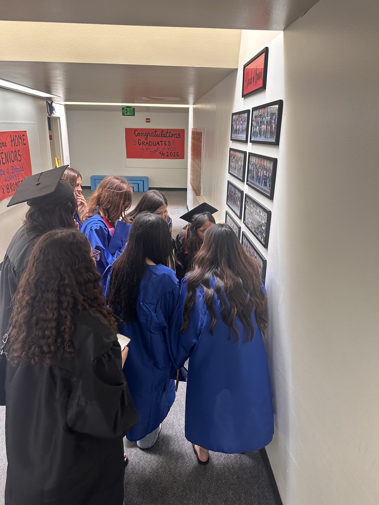 Graduating seniors in caps and gowns gather in a school hallway, looking at framed class photos on the wall