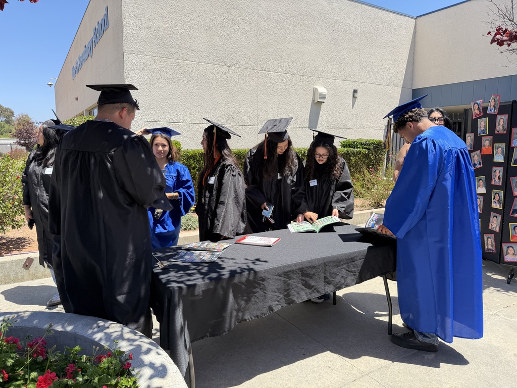 group of high school grad students in front of school wearing caps and gowns looking at yearbooks