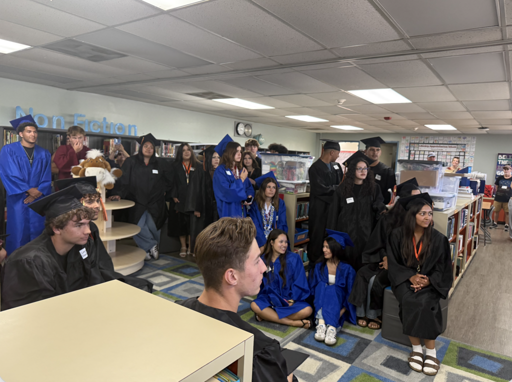 Graduating seniors in caps and gowns gather in the school library, listening and visiting during their campus walk.