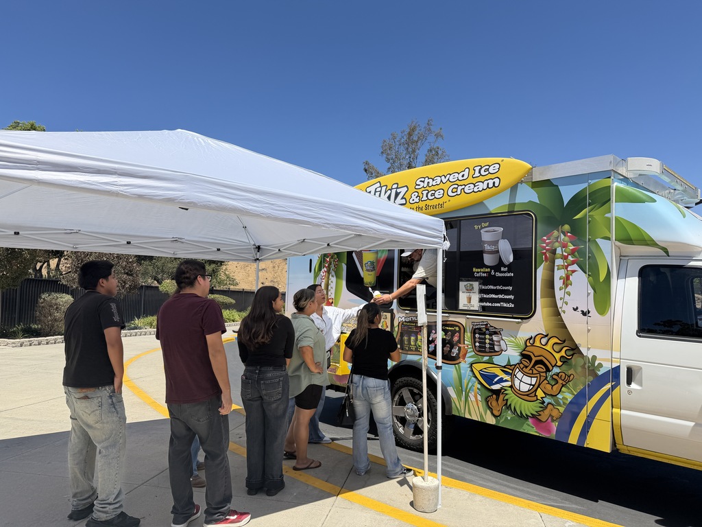 People queue under a white tent to order treats from a tropical-themed shaved ice and ice cream truck.