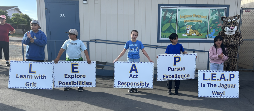 Children holding signs that spell out "L.E.A.P." at a school event, alongside a jaguar mascot. The signs include phrases like "Learn with Grit" and "Pursue Excellence."