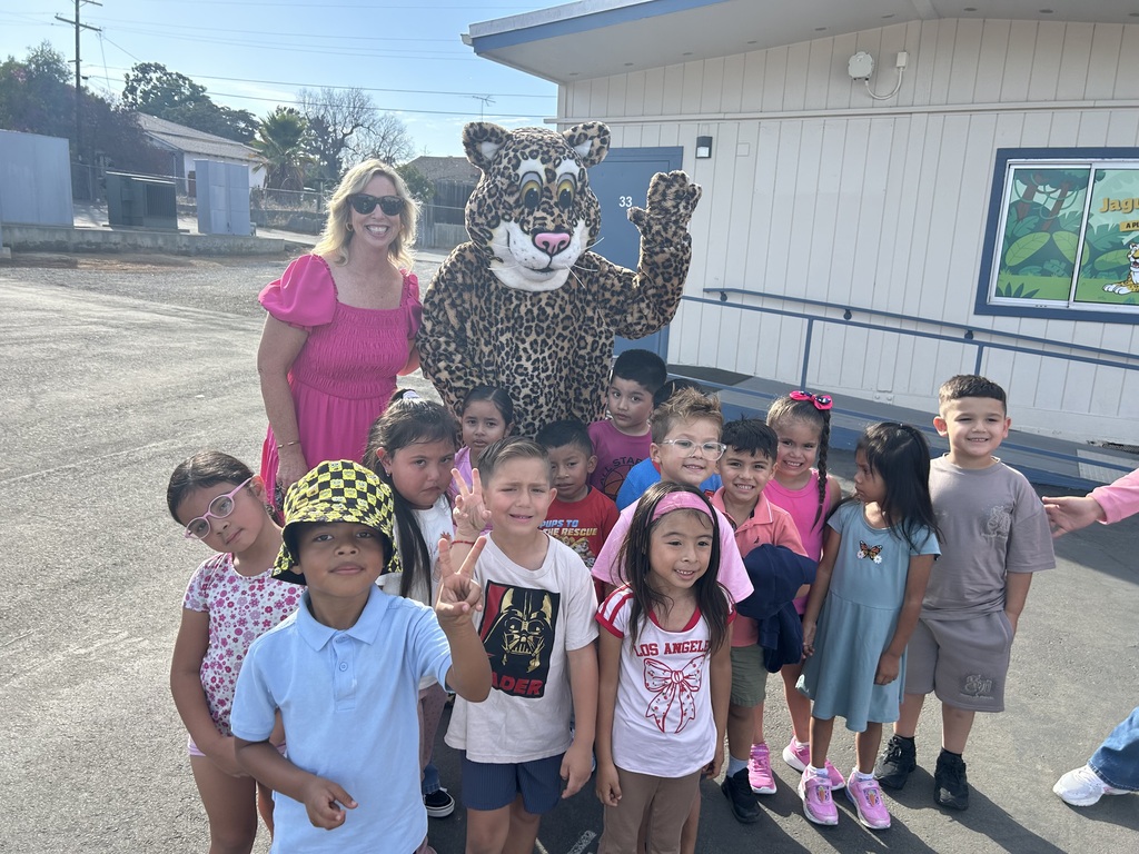A woman in a pink dress poses with a jaguar mascot and a group of smiling children outside a school on a sunny day