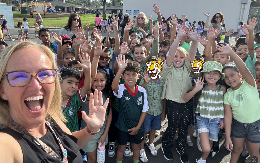 A group of Juniper Elementary students and staff smile and wave at the camera outside on campus