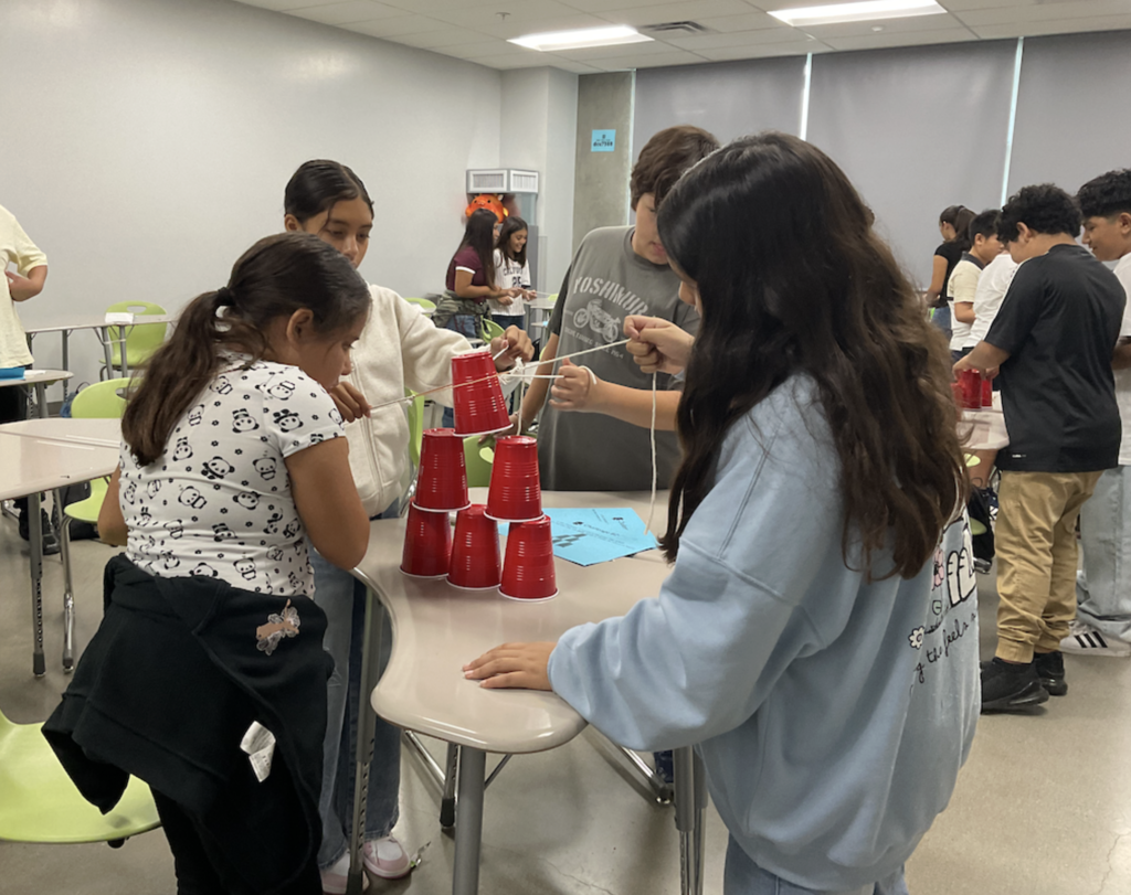 Dragon students pulling strings on a rubber band to stack cups during a teamwork exercise.