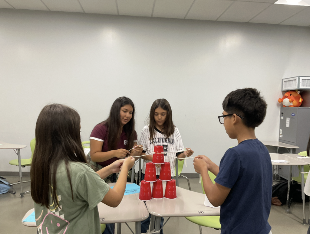 Dragon students pulling strings on a rubber band to stack cups during a teamwork exercise.