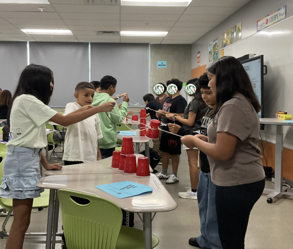 Dragon students pulling strings on a rubber band to stack cups during a teamwork exercise.