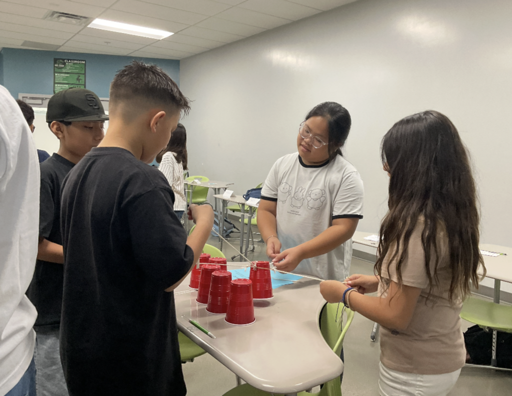 Dragon students pulling strings on a rubber band to stack cups during a teamwork exercise.