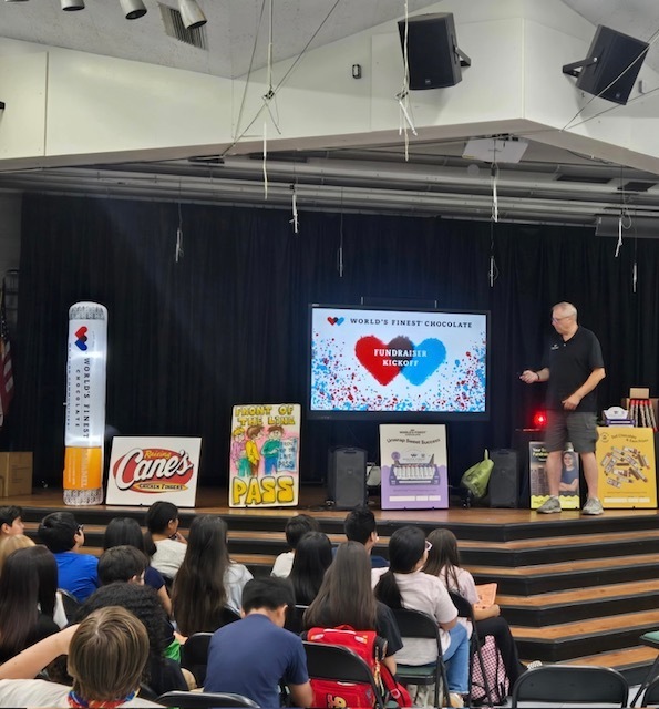 A presenter stands on a stage addressing an audience seated in folding chairs during a "World's Finest Chocolate" fundraiser kickoff. The stage features a large central digital screen displaying a graphic with red and blue hearts that reads "World's Finest Chocolate Fundraiser Kickoff."  To the left of the screen, an inflatable World's Finest Chocolate bar stands upright, accompanied by a sign for "Raising Cane's Chicken Fingers" and a colorful poster with the text "Front of the Line Pass." To the right of the screen, additional promotional posters are on display. The audience in the foreground is viewed from behind as they watch the presentation.