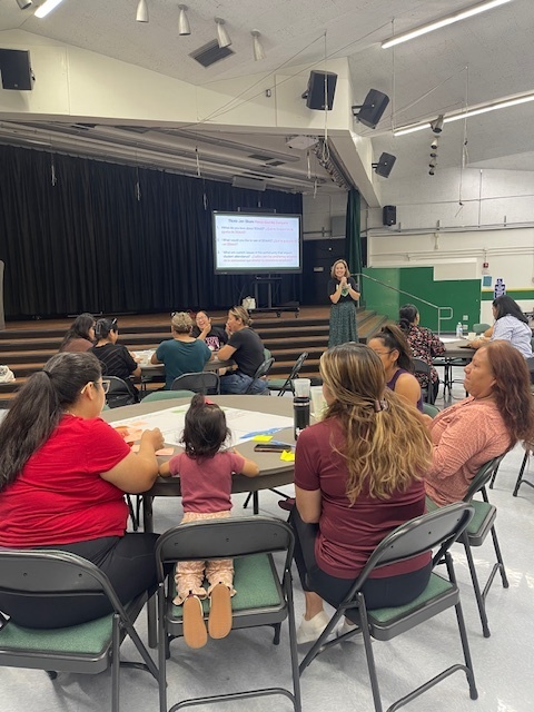 A group of adults and a young child are seated around tables in what appears to be a school auditorium or multipurpose room, participating in a workshop or presentation. A screen displaying a presentation slide is visible in the background near a stage.