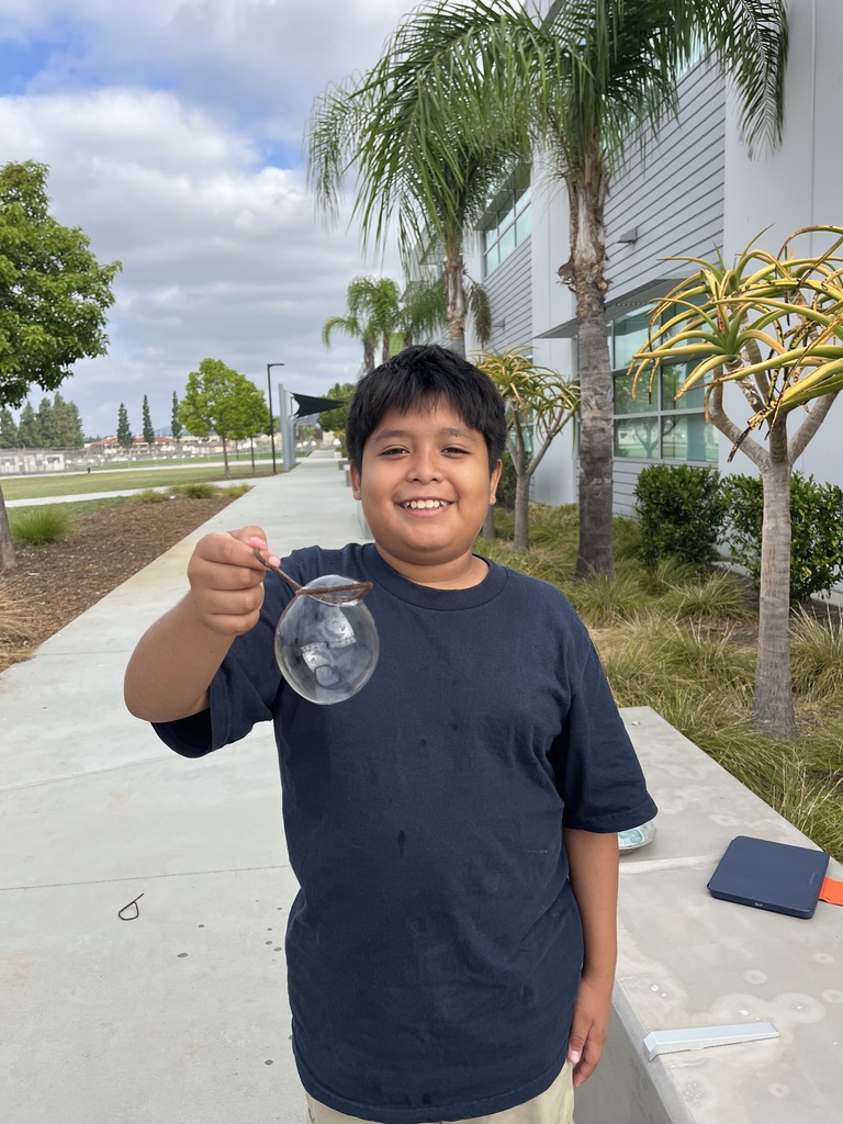 A student is taking part in a hands-on science activity to learn about how cell membranes work. He is holding a bubble wand with a bubble underneath it.  