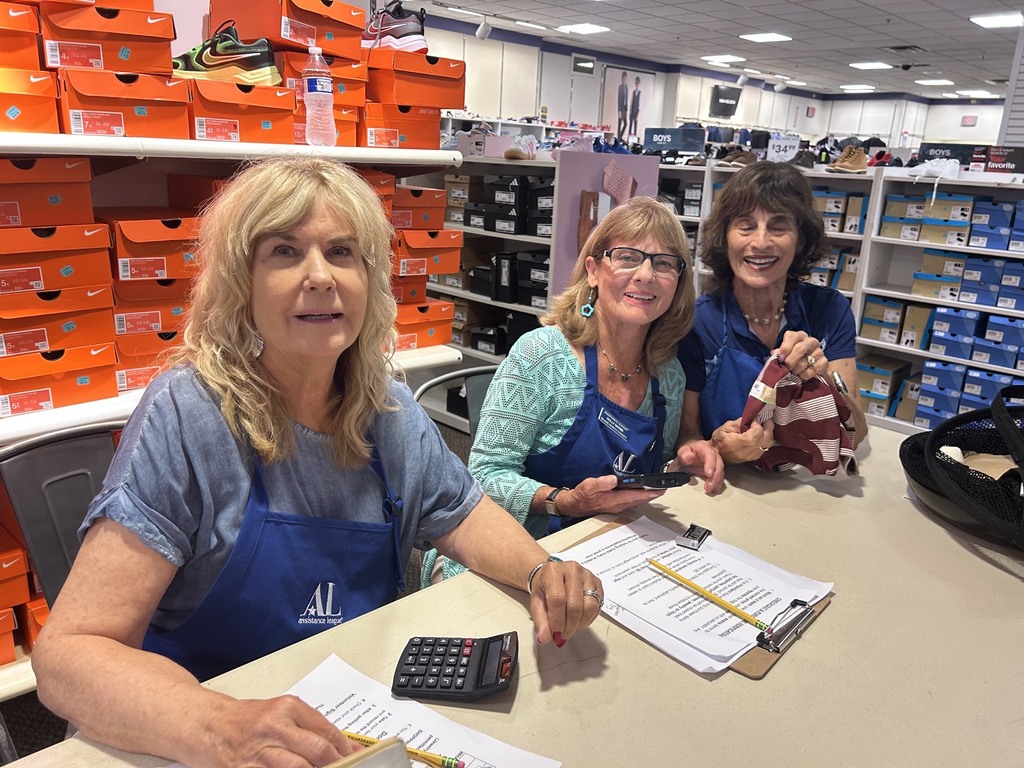 Three women in blue aprons pose behind a shoe-store counter surrounded by Nike shoeboxes, shoes, paperwork, and a calculator.