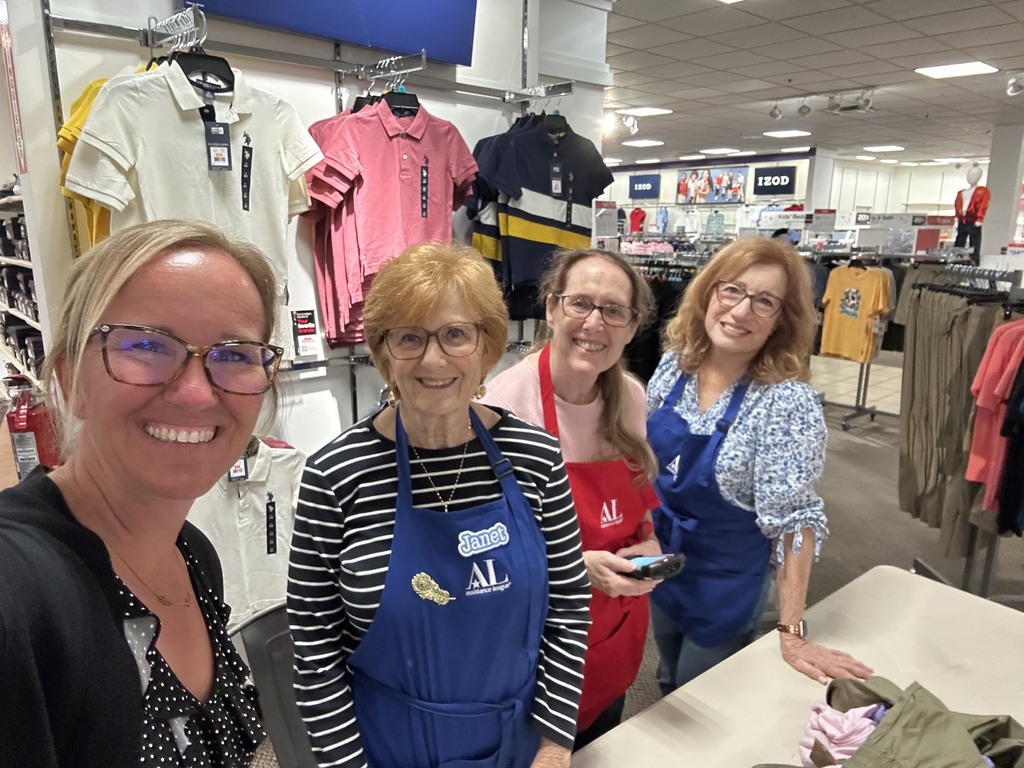 Four smiling women pose for a selfie inside a clothing store surrounded by racks of shirts and other apparel.