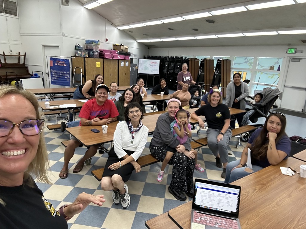 Group selfie of adults and children gathered around tables in a brightly lit classroom, with a laptop in the foreground and an EXIT sign in the background.