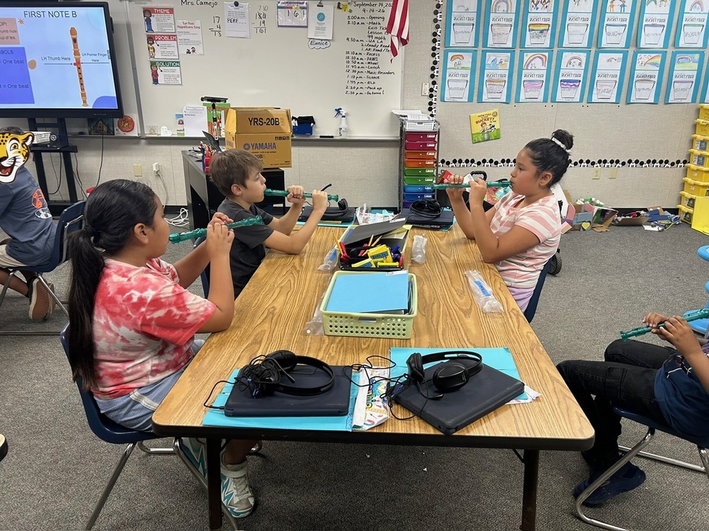 Students practice green recorders around a classroom table while a recorder fingering diagram and classroom displays are visible in the background.