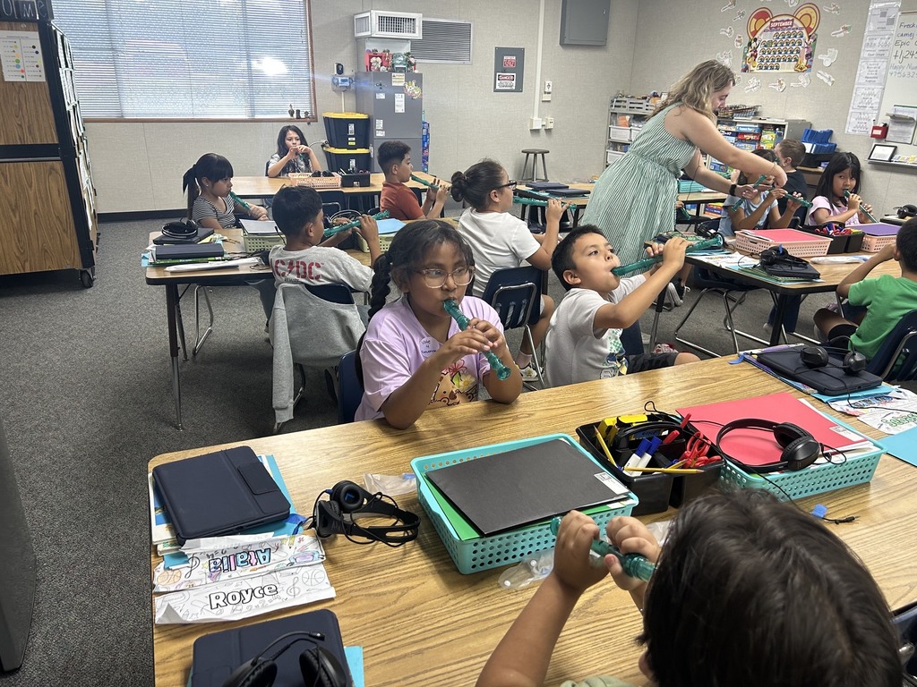 Teacher assists children playing recorders in a classroom filled with tables and school supplies.