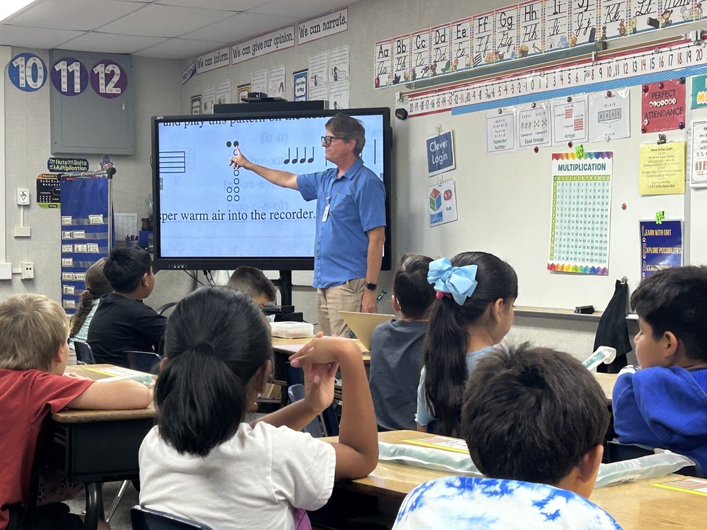Teacher points to musical symbols on an interactive display while students watch in a decorated classroom.