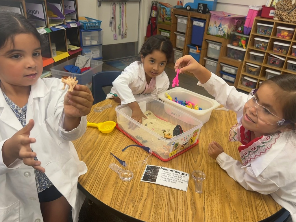 Children in lab coats explore a sensory bin and dinosaur-themed materials at a classroom table.