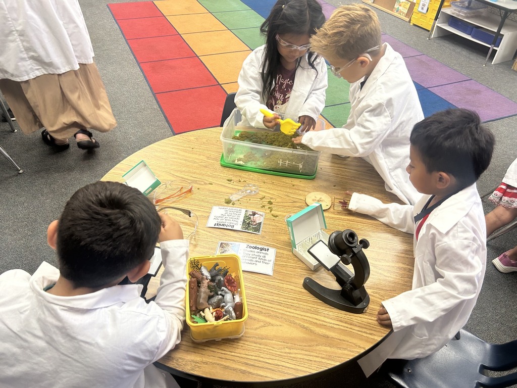 Children in white lab coats conduct a hands-on science activity with moss, toy animals, and a microscope at a classroom table.