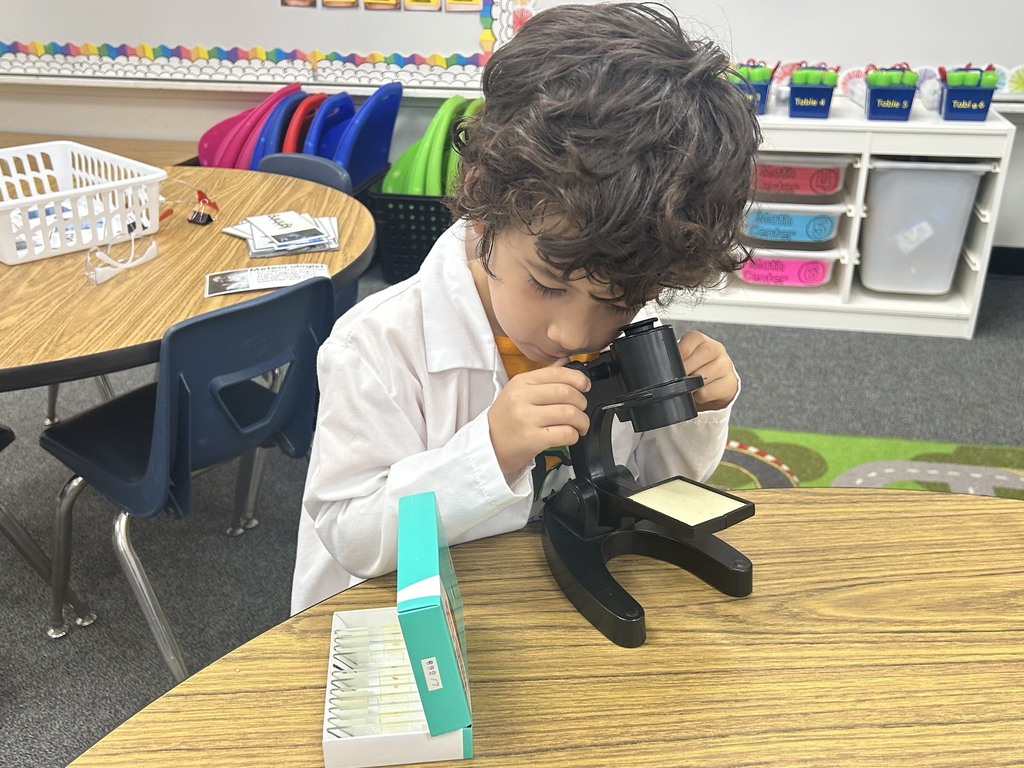 Child in a white lab coat examining a specimen through a microscope in a classroom.