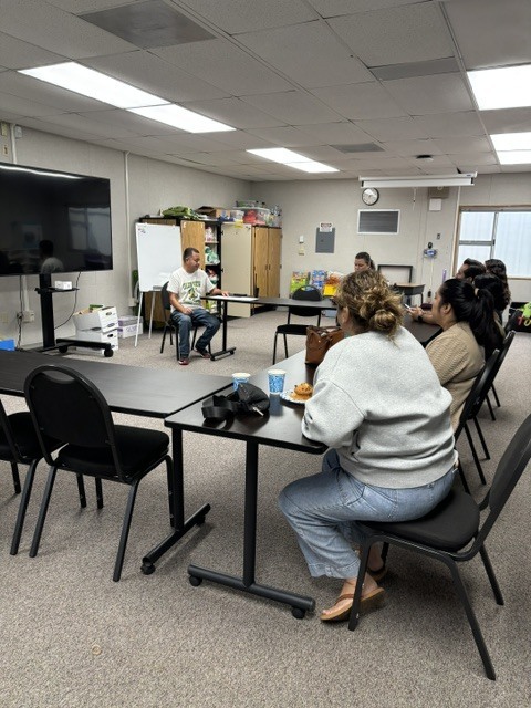 Four parents are sitting on black chairs and using black and brown tables as they listen to Principal Lopez o the right hand side of the room. 