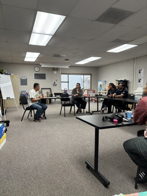 Principal Lopez is sitting on the left next to a white board and is speaking to four parents inside room 21. 