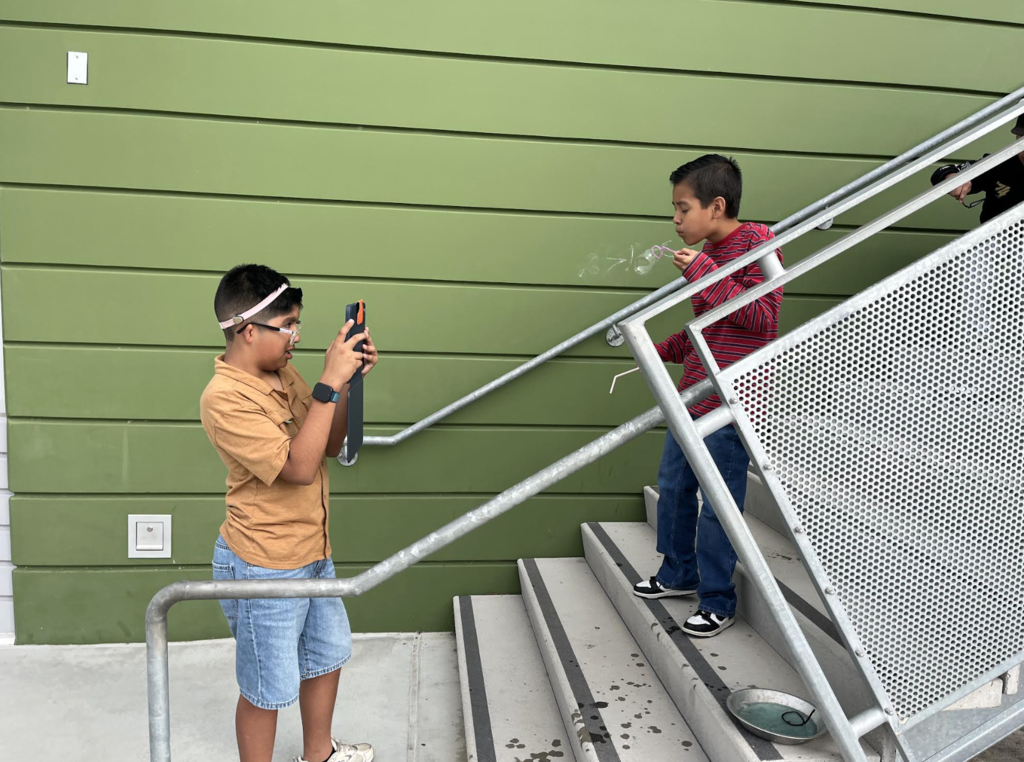 Two students are taking part in a hands-on science activity to learn about how cell membranes work.  The student in red is blowing a bubble while the other is taking a picture of his classmate. 