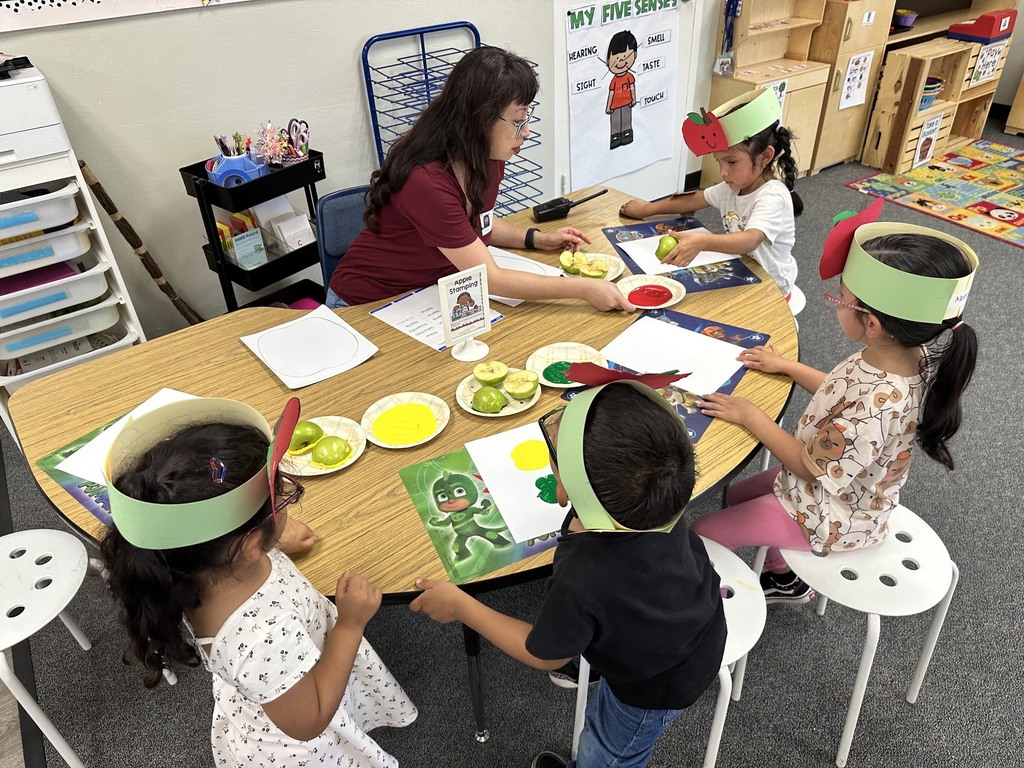 Children wearing apple crowns participate in an apple-themed classroom activity involving apple slices, worksheets.
