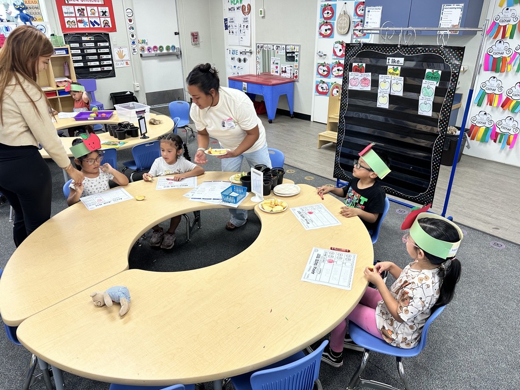 Children wearing apple crowns participate in an apple-themed classroom activity involving apple slices, worksheets.