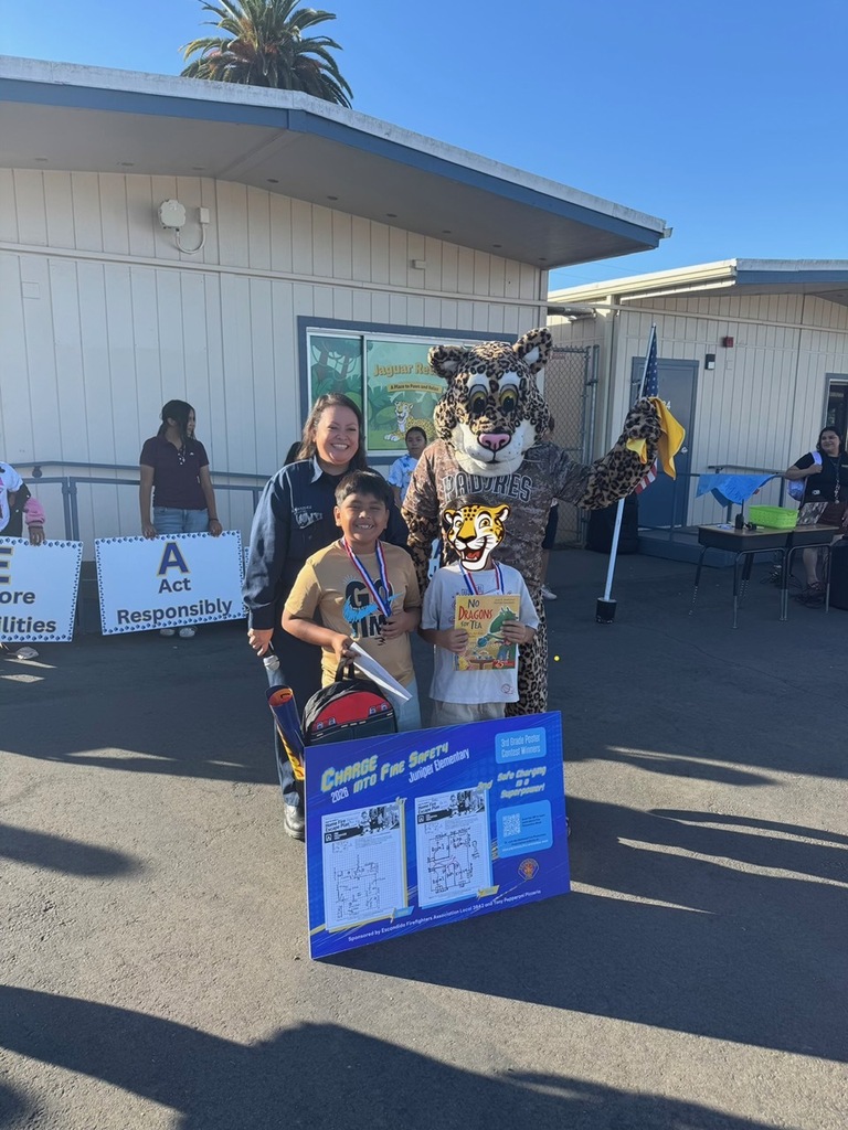 Children, an adult, and a jaguar mascot pose outside a school while holding a fire-safety poster and a children’s book.