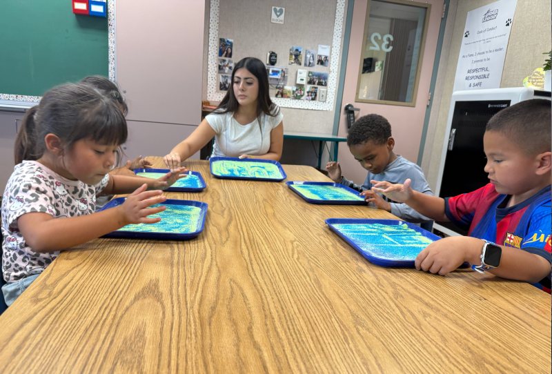 Kindergarten students sit at a table with a teacher and play with glitter.