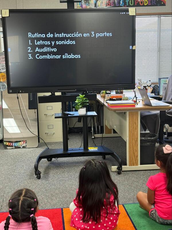 Kindergarten students sit on a floor and view a television stating, "Rutina de instruccion en 3 parte: 1. Letras y sonidos, 2. Auditivio, 3. Combinar silabas.