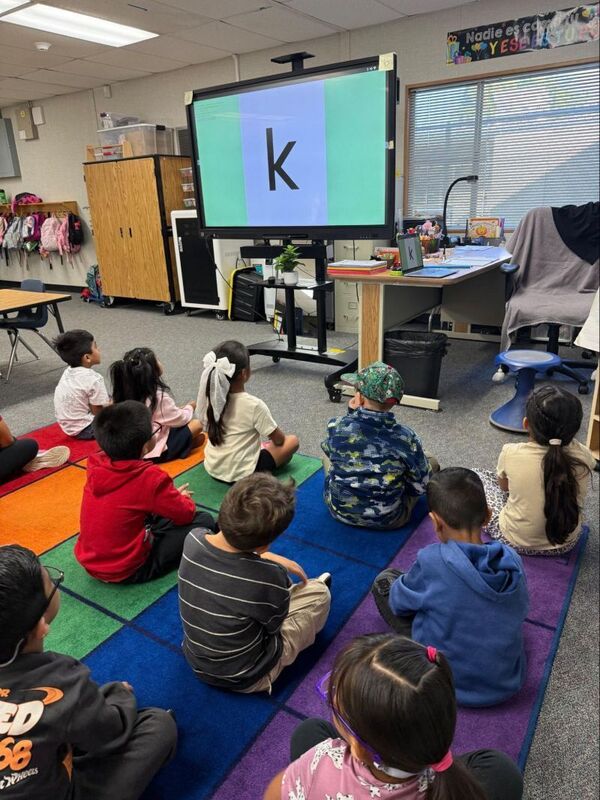 Kindergarten students sit on the floor and learn language pattern recognition, with the letter "k" on a television.