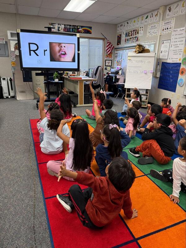 Students sit on the floor of their classroom and view a television with the letter "R."