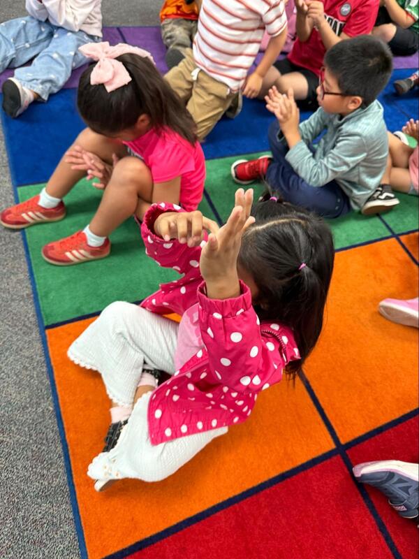 Students sit on the floor of a classroom and raise hands.