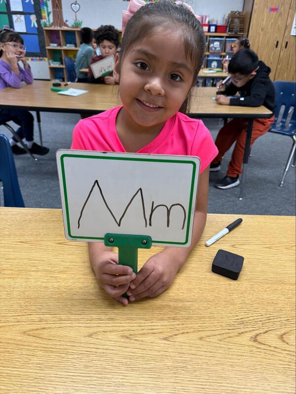 A student holds a white board with the letter "Mm."