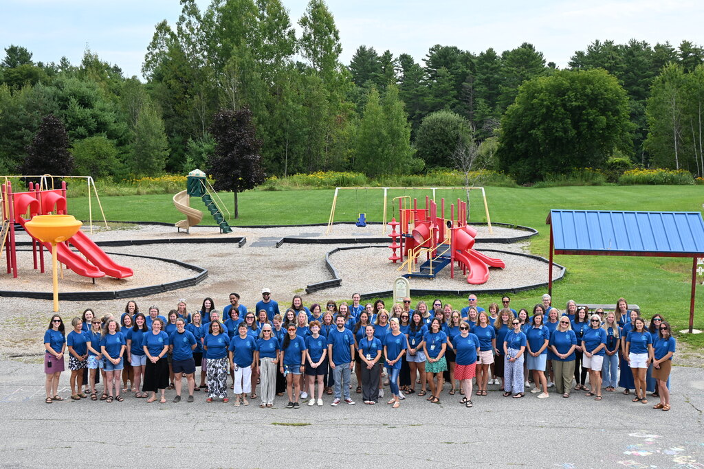 A group of people in blue shirts stands in front of a playground with slides and trees.