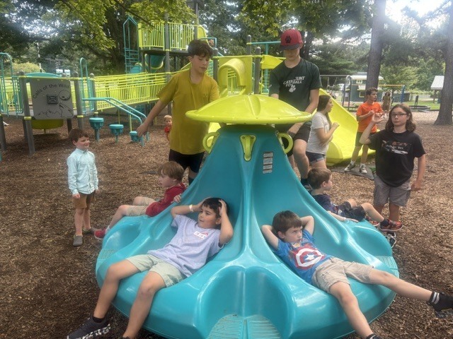 Several children in a park, with some on a blue and yellow spinning ride, and others standing around.