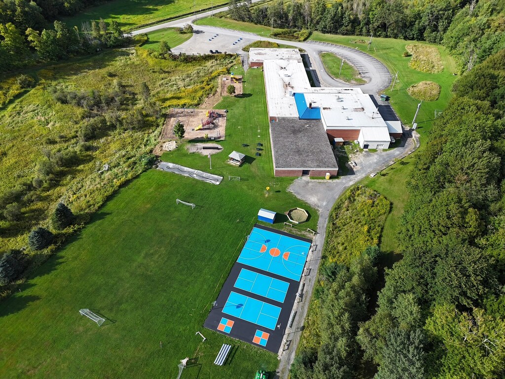 Aerial view of a large building with a blue swimming pool. The building is near a green field and trees.